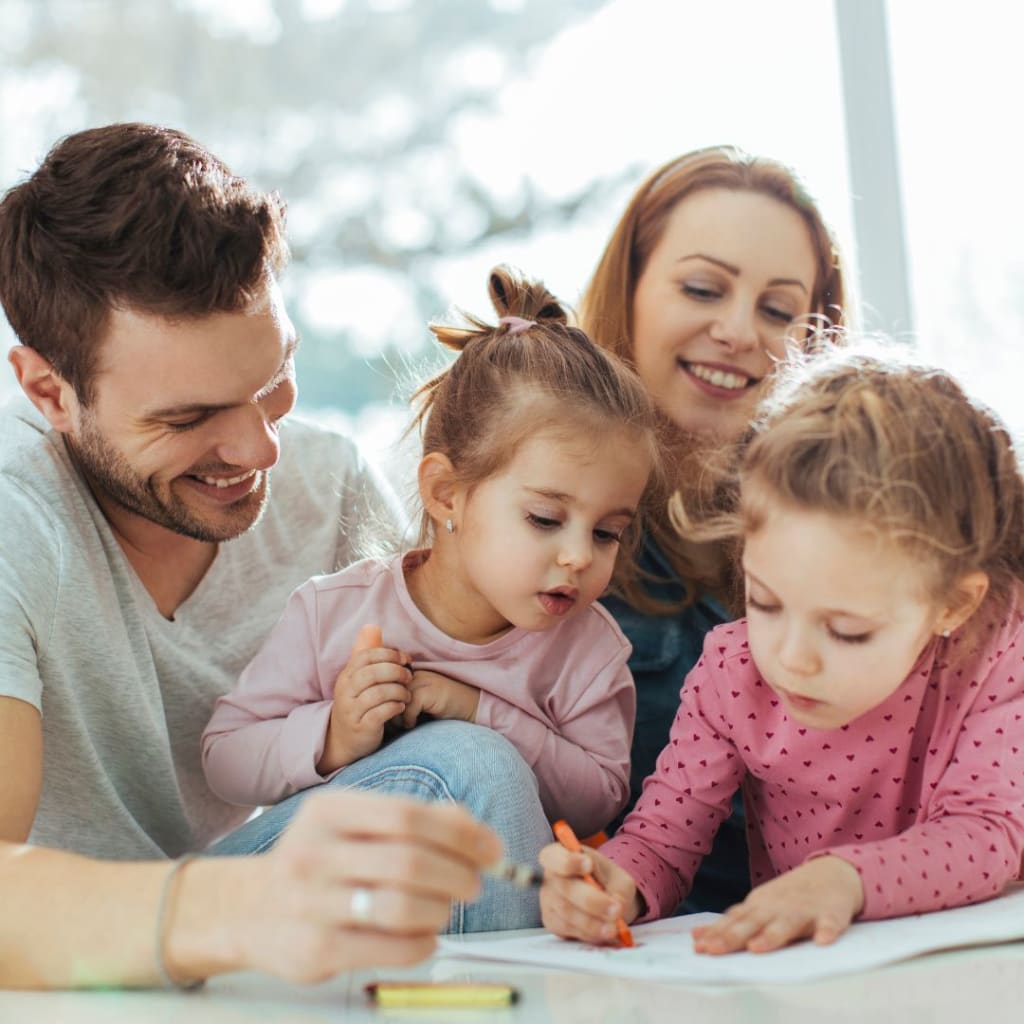 A family sits together. Two young children are drawing while a man and a woman watch and smile. The setting is well-lit and cheerful.